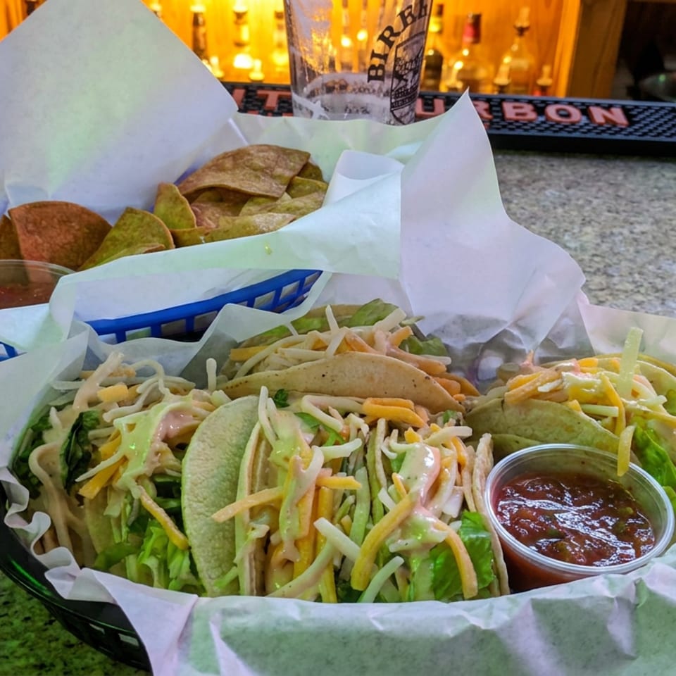 Basket of soft tacos topped with shredded lettuce, cheese, and sauce beside tortilla chips and salsa at a bar.